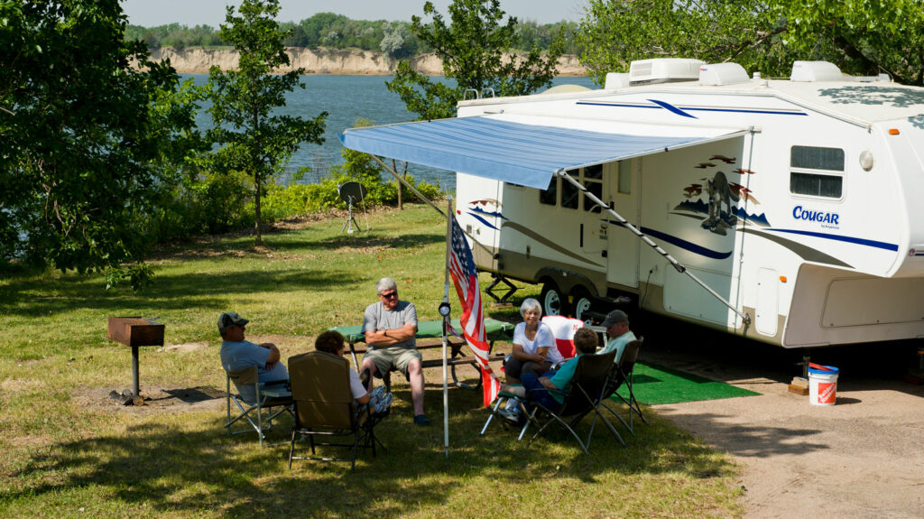 people sitting in a circle in camp chairs in the shade next to an RV