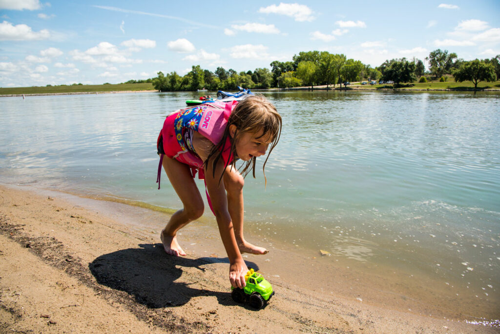 A young girl wearing a lifejacket plays with a car on the beach.