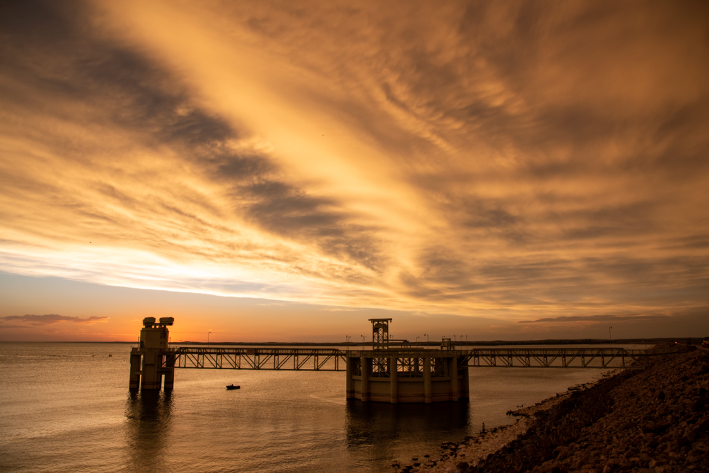 The sun sets behind an outlet structure on a lake