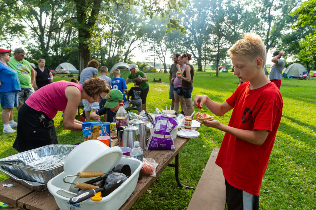 many people gather to eat camp food near the fire and picnic table