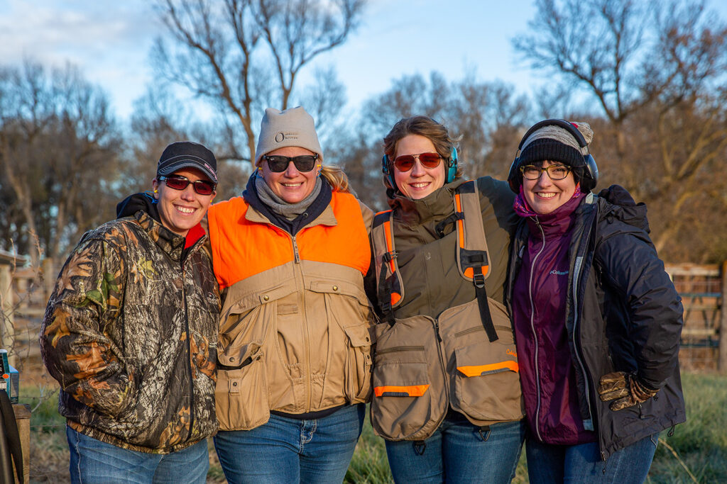 Four women pose and smile in hunting gear while standing outside