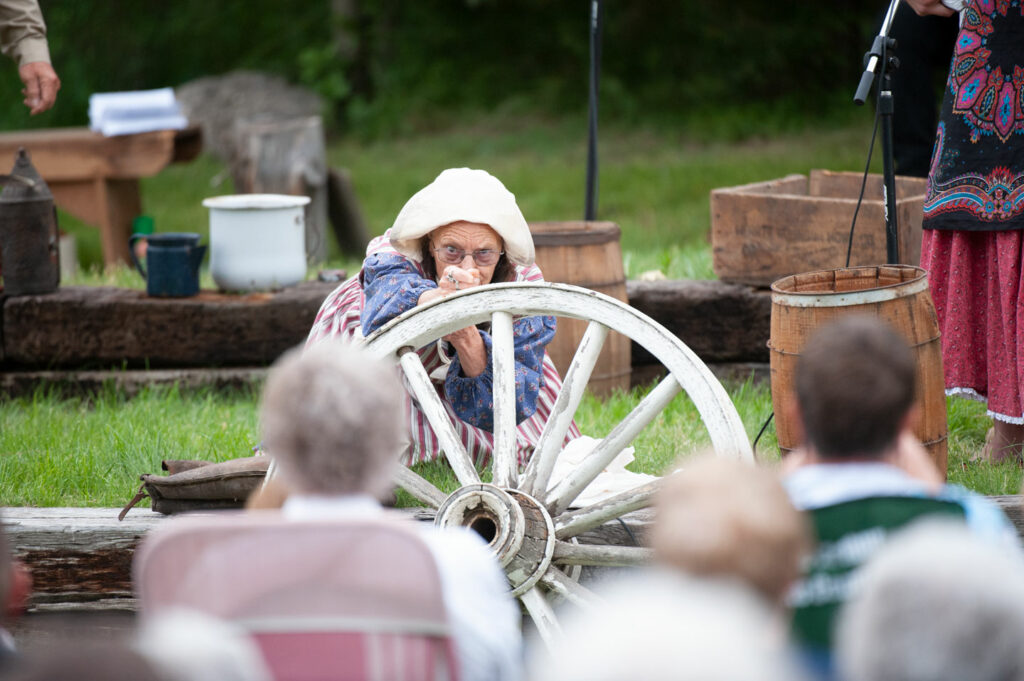 A woman peers over a wagon wheel painted white, performing in a play.