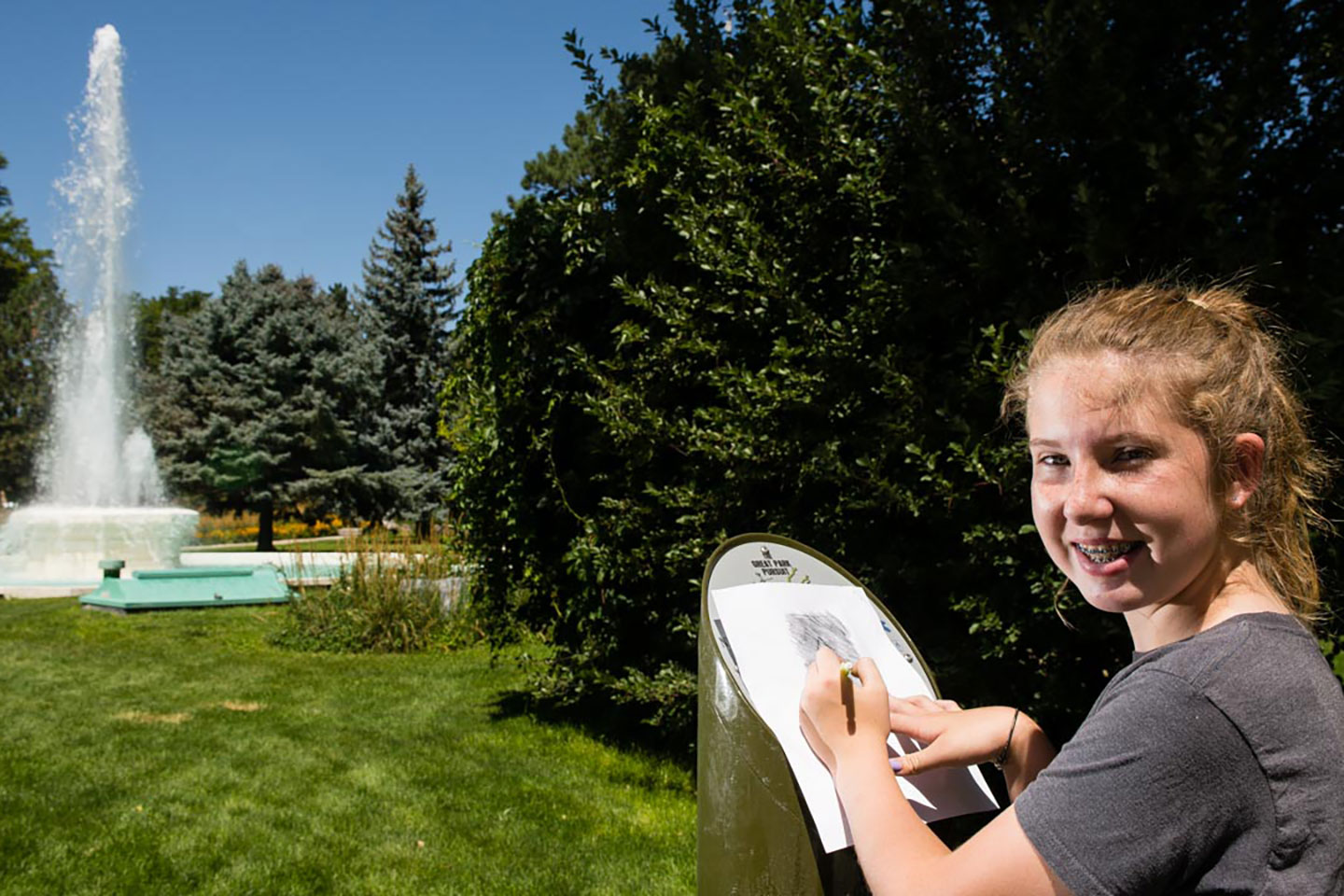 A girl is tracing (rubbing) with a pencil over the Great Park Pursuit post at a park in Alliance City Park. A water fountain in the background.