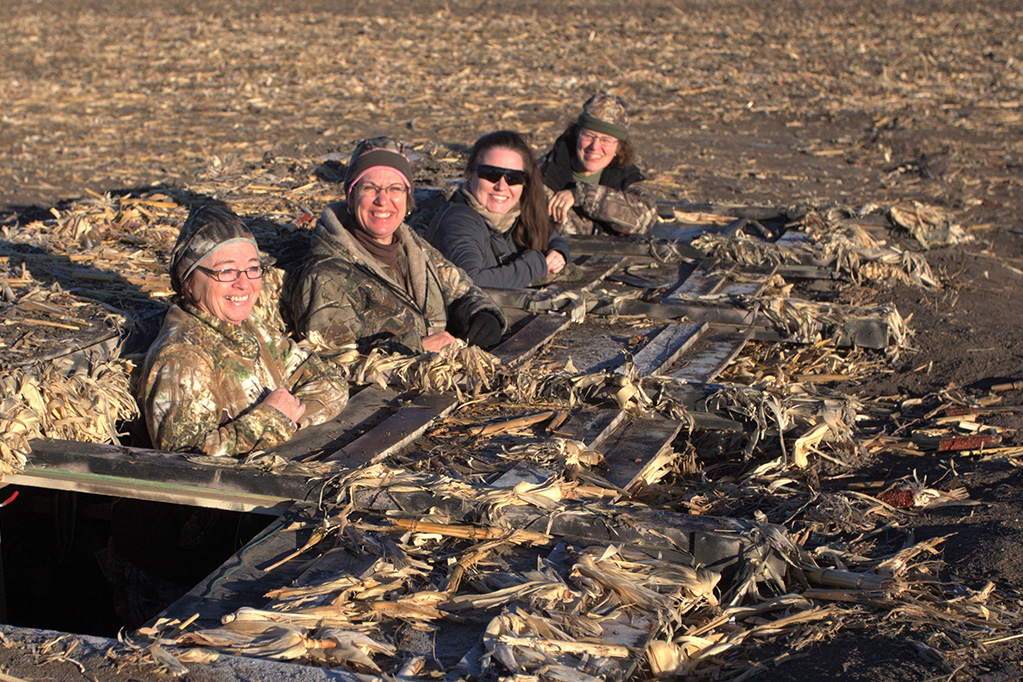 Four smiling ladies sit in a duck hunting blind in a Nebraska cornfield