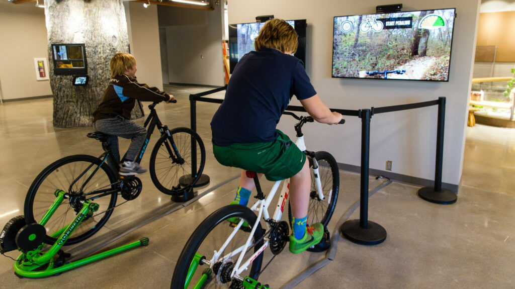 two people ride stationary bikes that are connected to televisions showing a virtual mountain bike trail