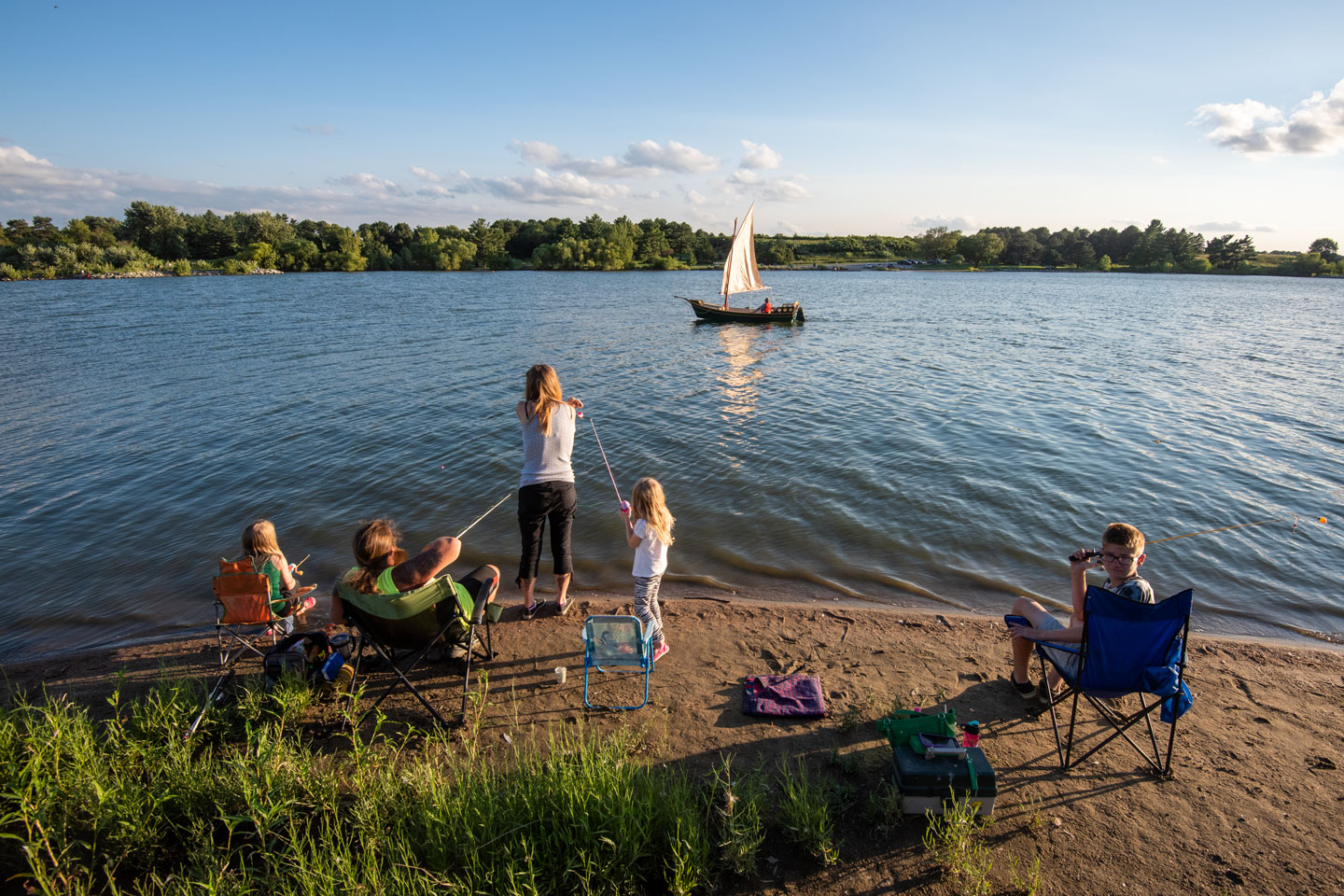 Five family members fish on the bank of a lake as a sailboat passes on a clear summer day.