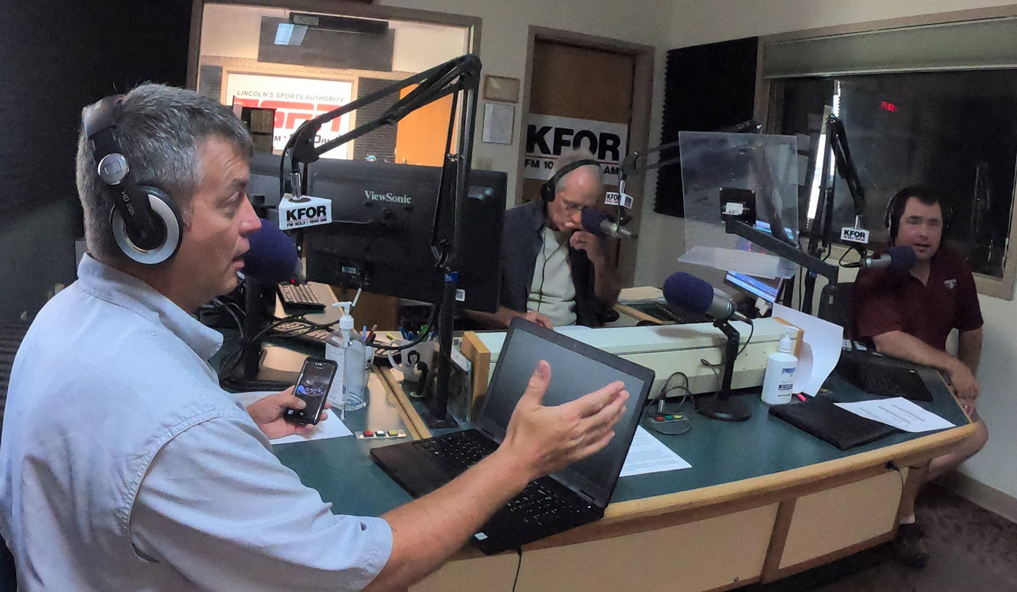 Three men sit in a radio studio wearing audio equipment.