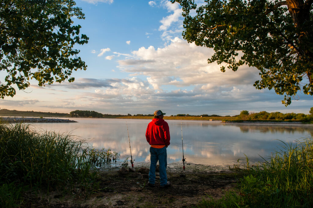 A man stands in the foreground fishing at a scenic lake with two rods planted on the shoreline