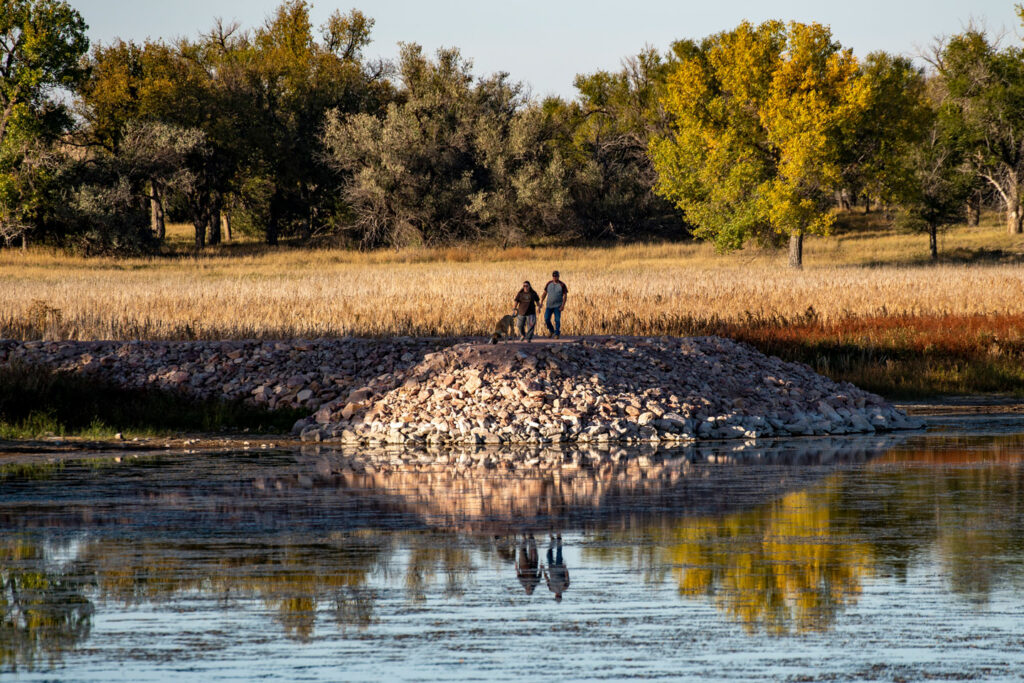 Two people walk a dog on a rocky fishing pier at a lake during autumn